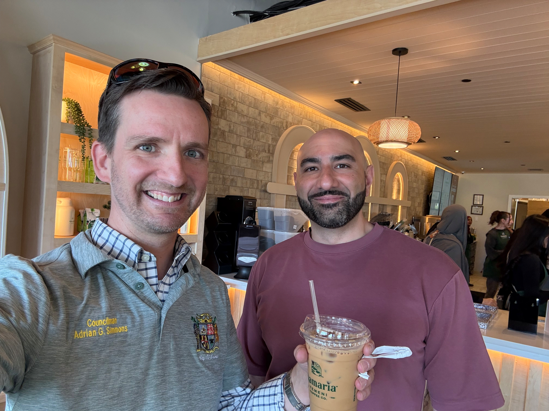 Auto-generated description: Two people are standing indoors, with one holding a cup of iced coffee in a cafe setting.