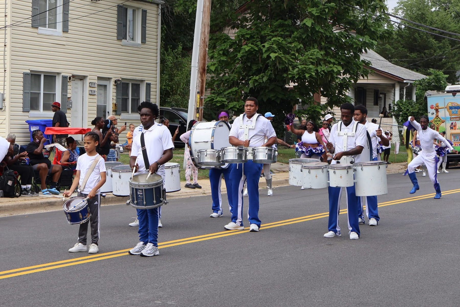 Auto-generated description: A marching band is performing in a parade on a residential street with onlookers watching from the sidewalk.
