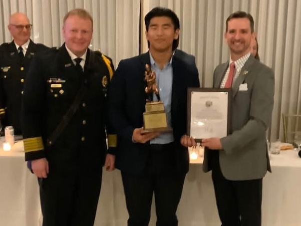 Auto-generated description: A young man is being presented with a trophy and a certificate by two men, one in a police uniform and the other in a suit, during an indoor event.