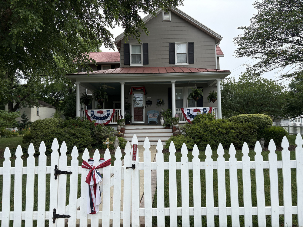 A two-story house with a red roof and a front porch decorated with patriotic bunting is viewed through a white picket fence adorned with a matching ribbon.