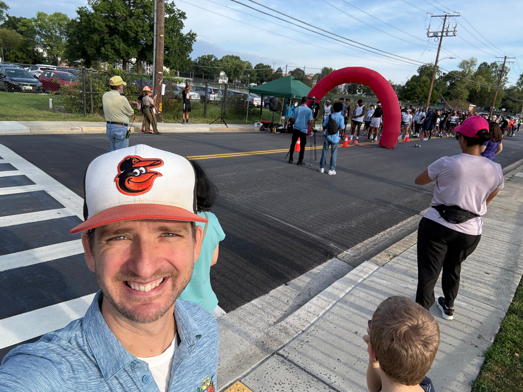 Auto-generated description: A man wearing a baseball cap with an Orioles logo is smiling and taking a selfie near a street where a group of people are gathered around a red inflatable arch.