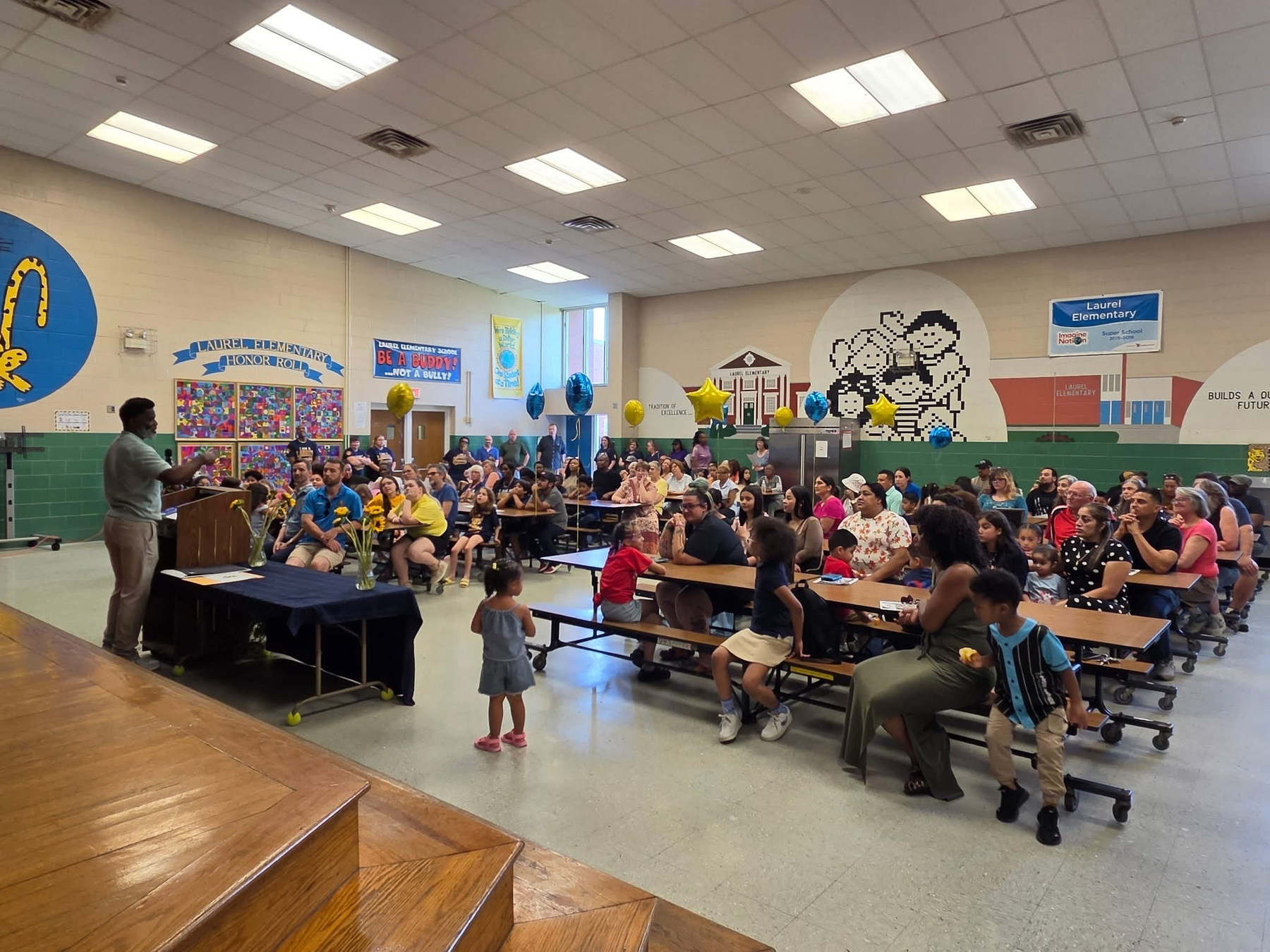 Auto-generated description: A speaker is addressing a large group of adults and children seated at cafeteria tables in a school multipurpose room decorated with colorful murals and banners.