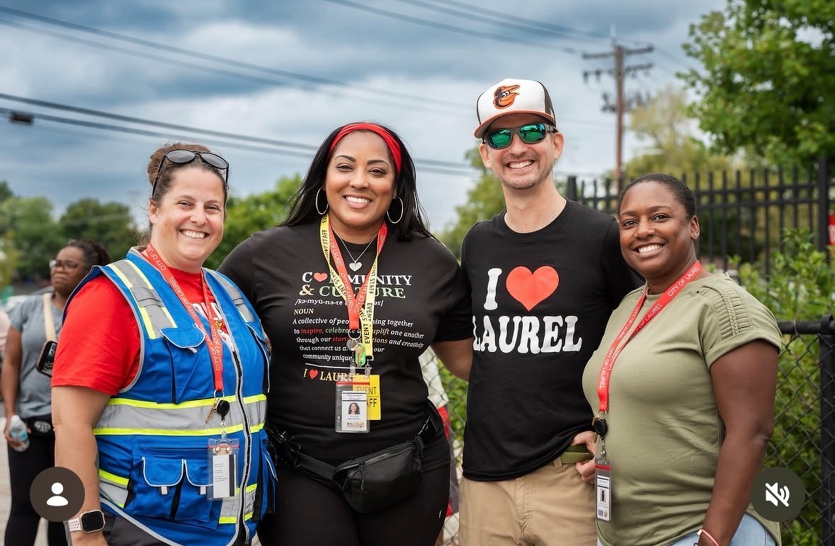 Auto-generated description: Four smiling people stand together outdoors, with two wearing lanyards and one wearing a shirt that says I ♥ LAUREL.