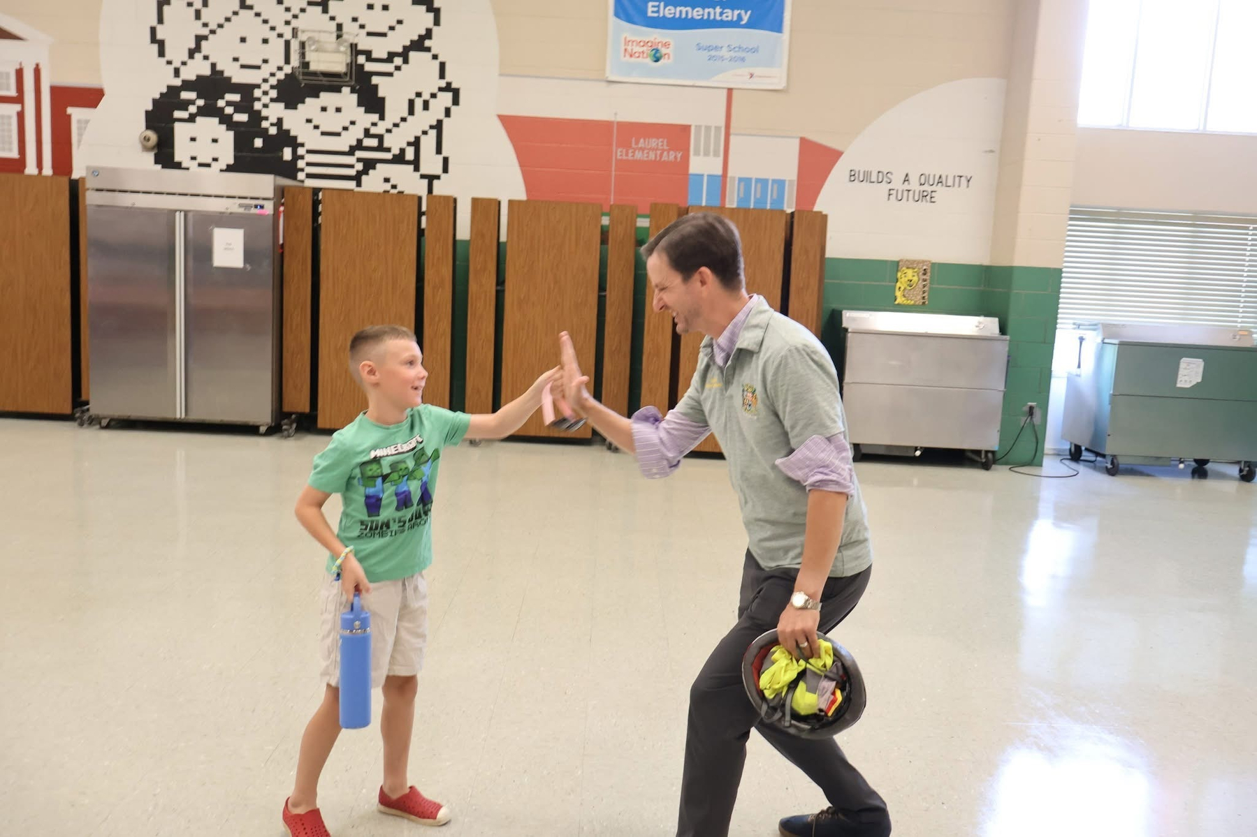 Auto-generated description: A young boy and a man are giving each other a high-five in a spacious indoor setting, possibly a school gym or cafeteria.