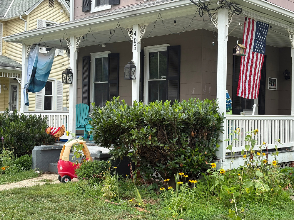 A cozy porch decorated with an American flag, outdoor chairs, lanterns, and children's toys overlooks a garden with blooming flowers and greenery.