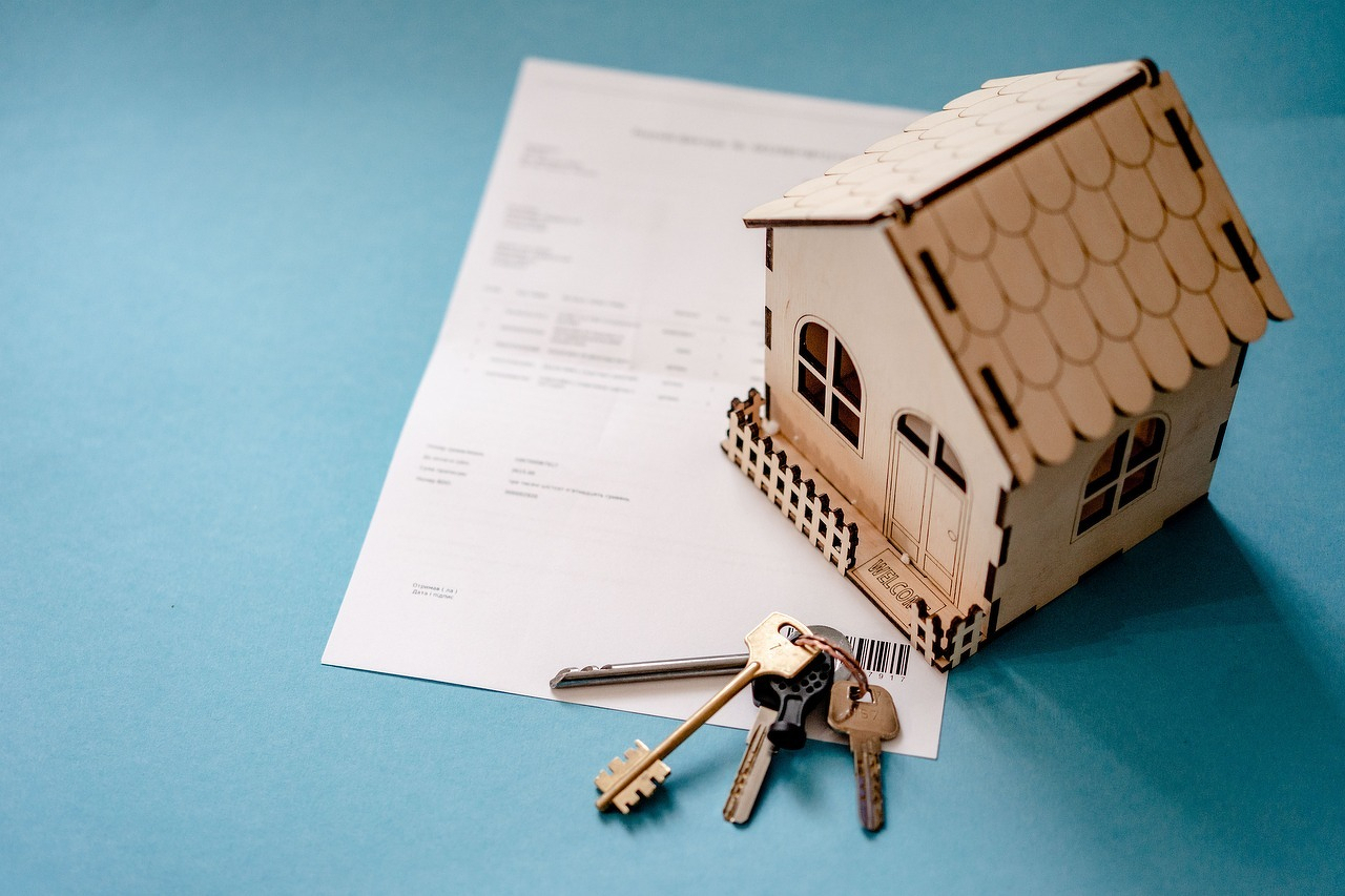 Auto-generated description: A small wooden model house sits on a document next to a set of keys on a blue surface.