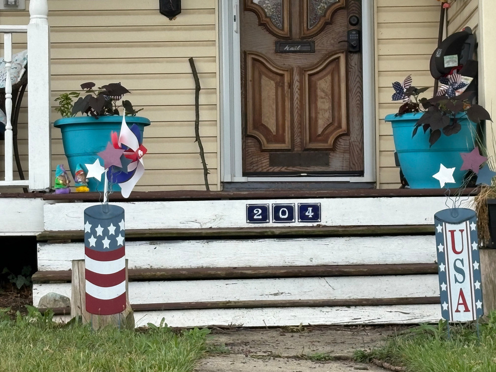 A porch decorated with two large blue flower pots, patriotic-themed wind spinners, and a house number 204 on the steps.