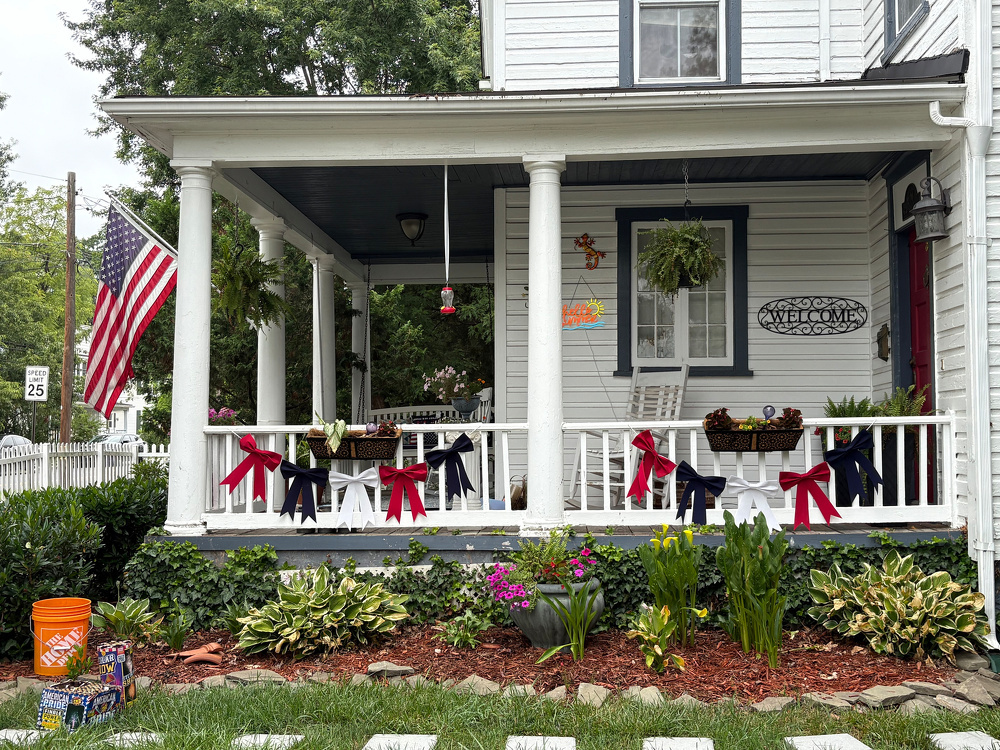 A charming porch decorated with red, white, and blue bows, hanging plants, an American flag, and a Welcome sign is surrounded by a well-maintained garden.