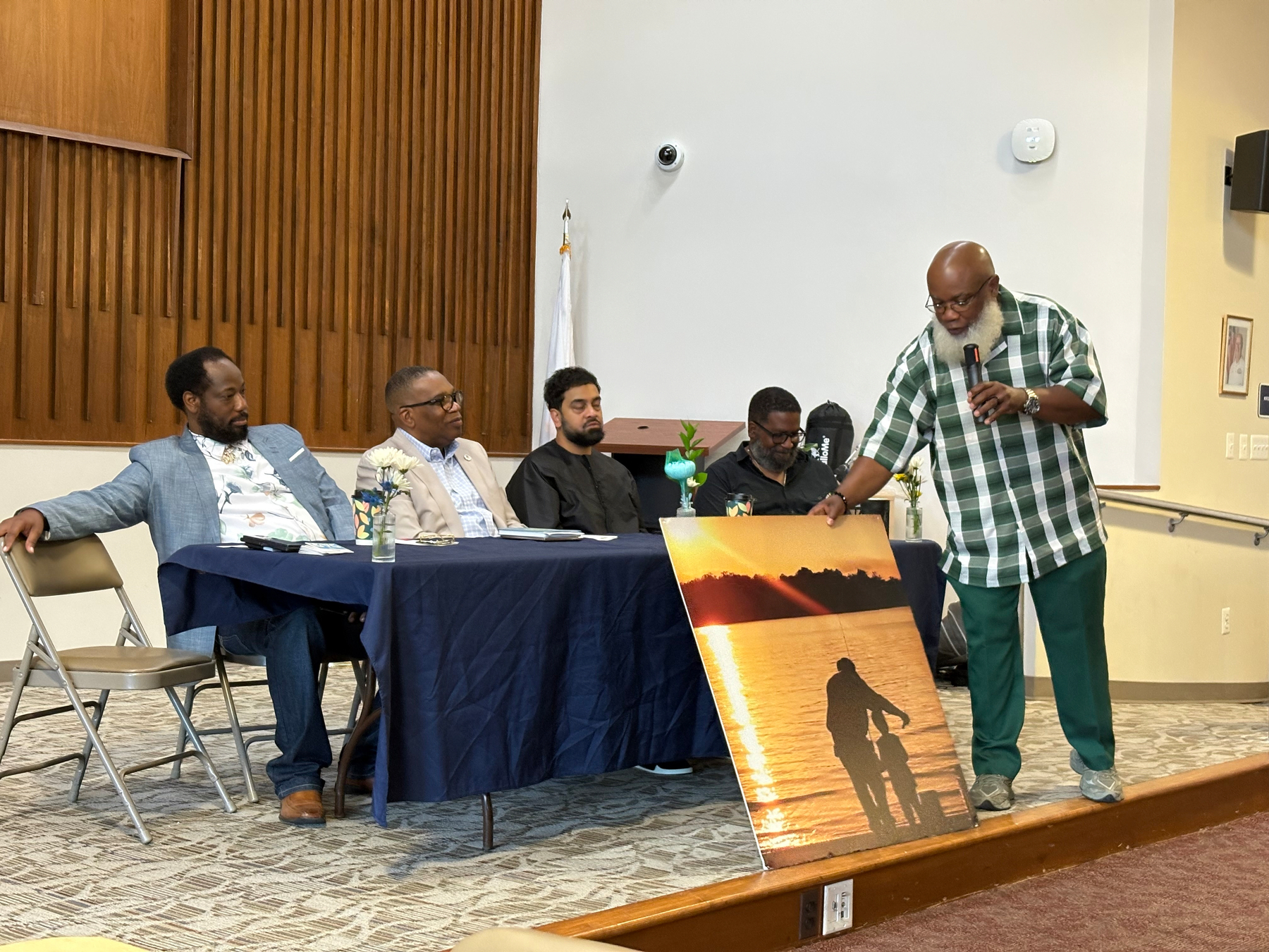 Auto-generated description: A man in a green plaid shirt is speaking into a microphone while pointing at a large photo of a person with a bicycle at sunset, with four other men seated at a table nearby listening.