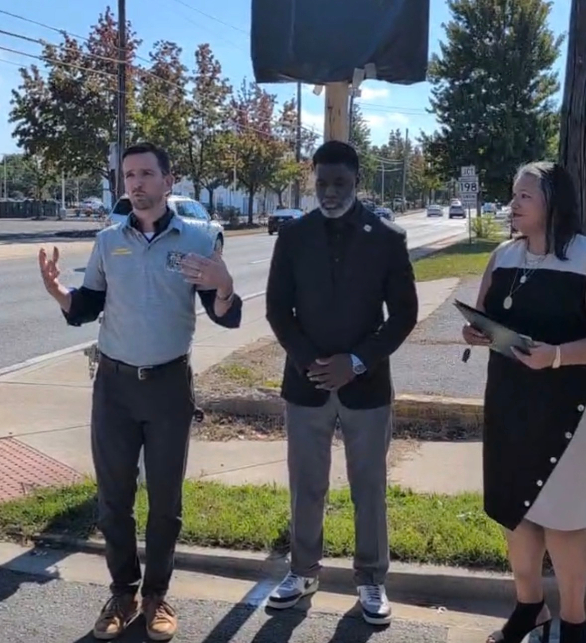 Auto-generated description: Three individuals are standing on a sidewalk near a street, with one man speaking and gesturing while the other two listen attentively.