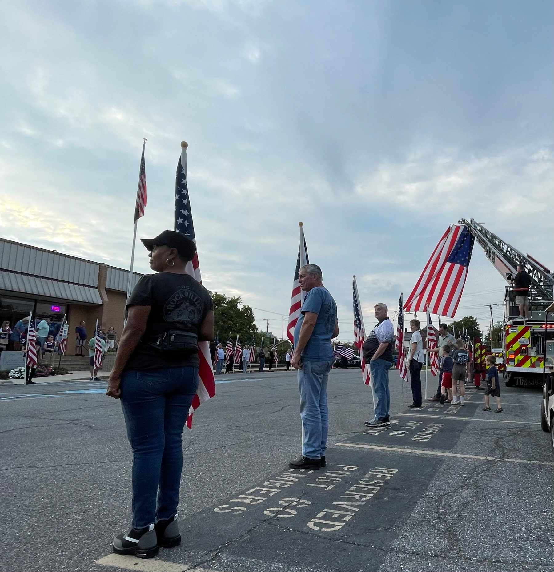 Auto-generated description: People stand in a line holding American flags along a street with a fire truck displaying a large American flag raised on a ladder.