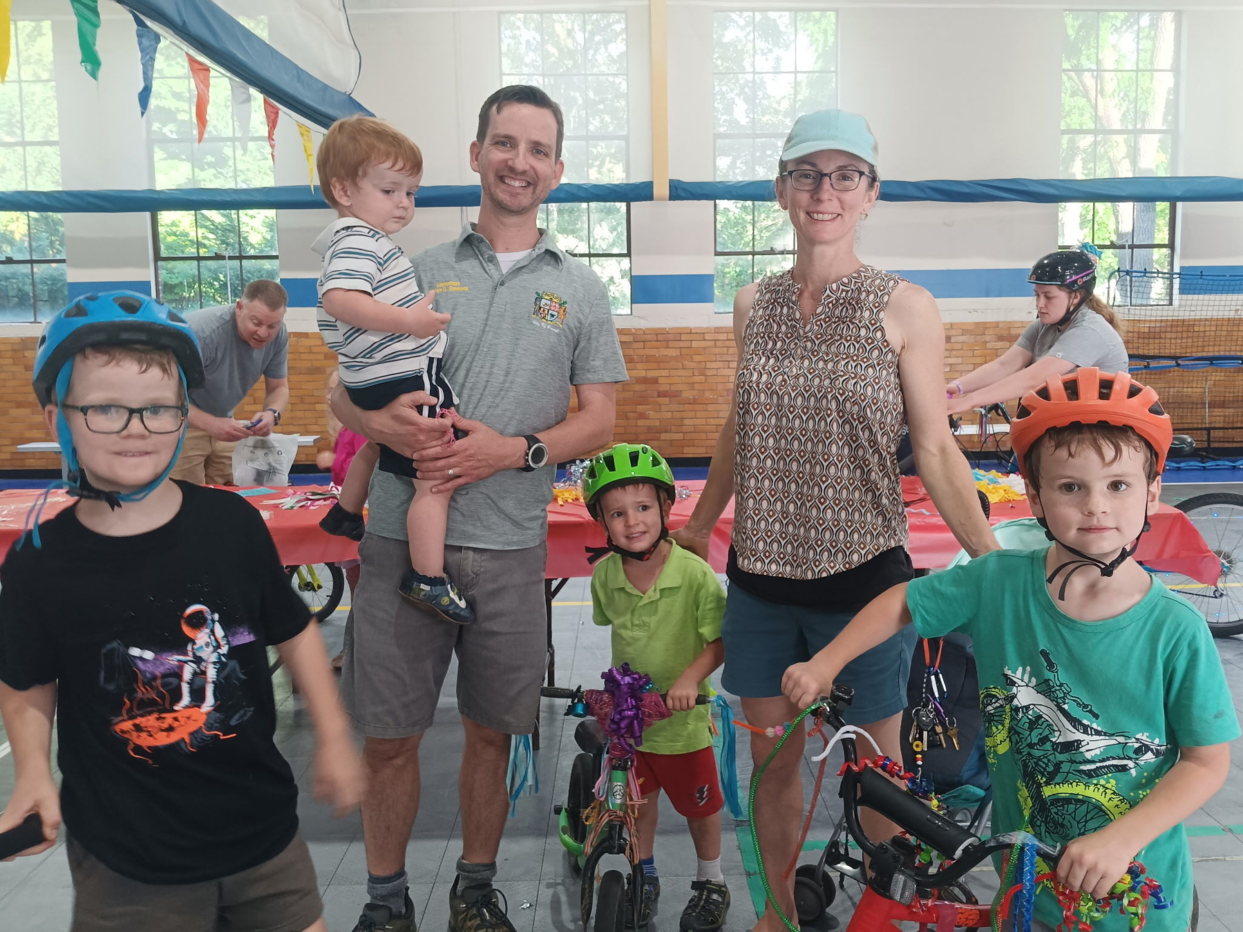 Auto-generated description: A family with three children wearing helmets and bicycles is posing indoors while a man and a woman stand behind them smiling.