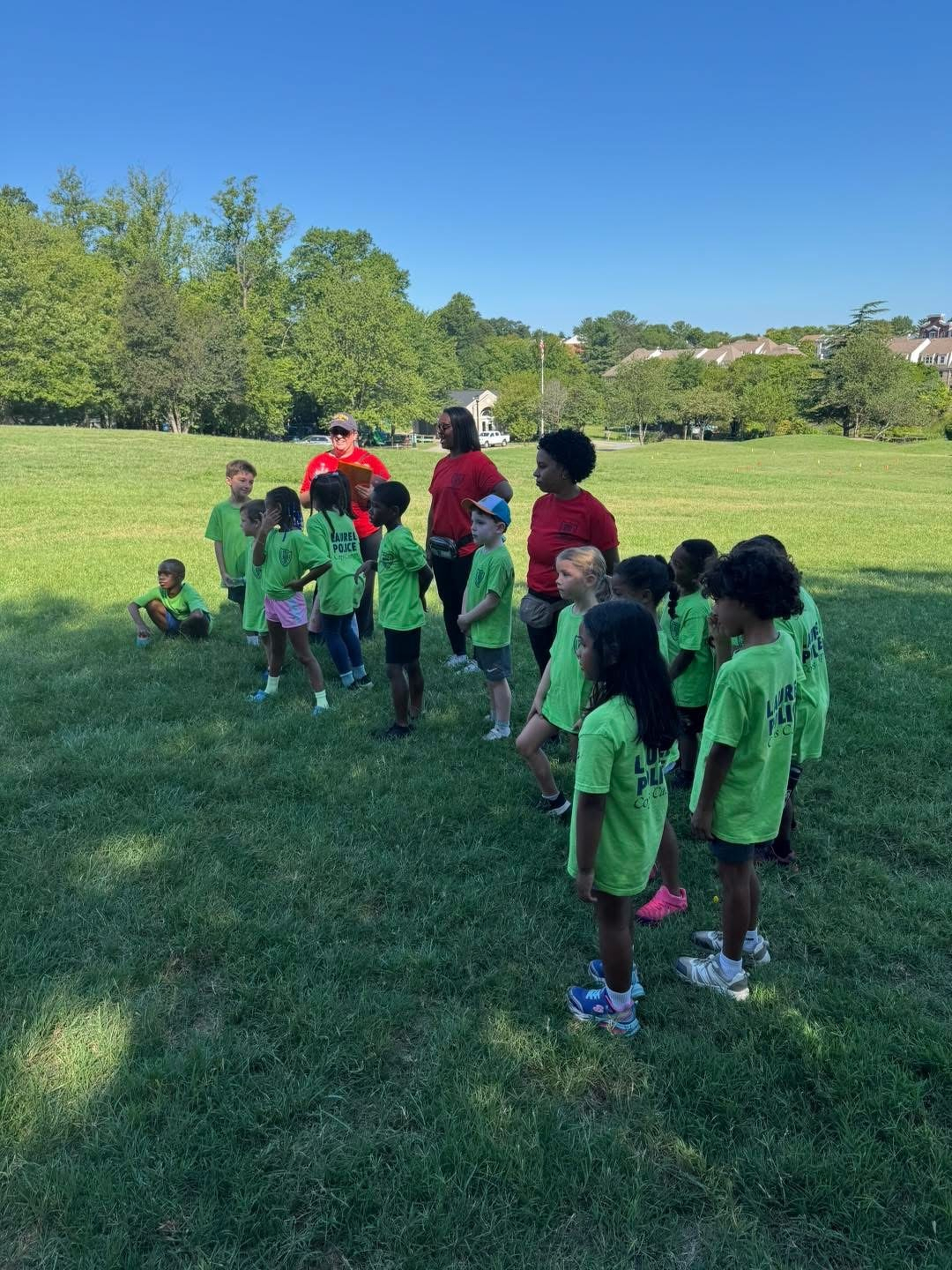 Auto-generated description: A group of children wearing matching green shirts are standing on a grassy field with a few adults in red shirts, seemingly participating in an outdoor activity or event.