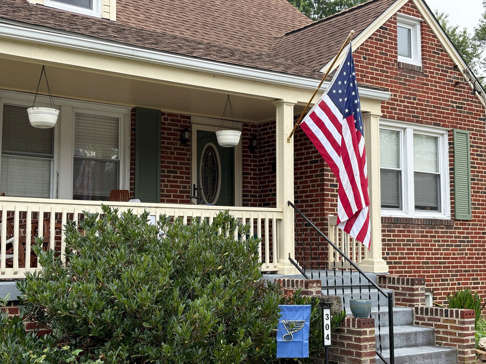 A red brick house features a white porch railing, an American flag displayed on a flagpole, and a prominent green shrub in the front yard.