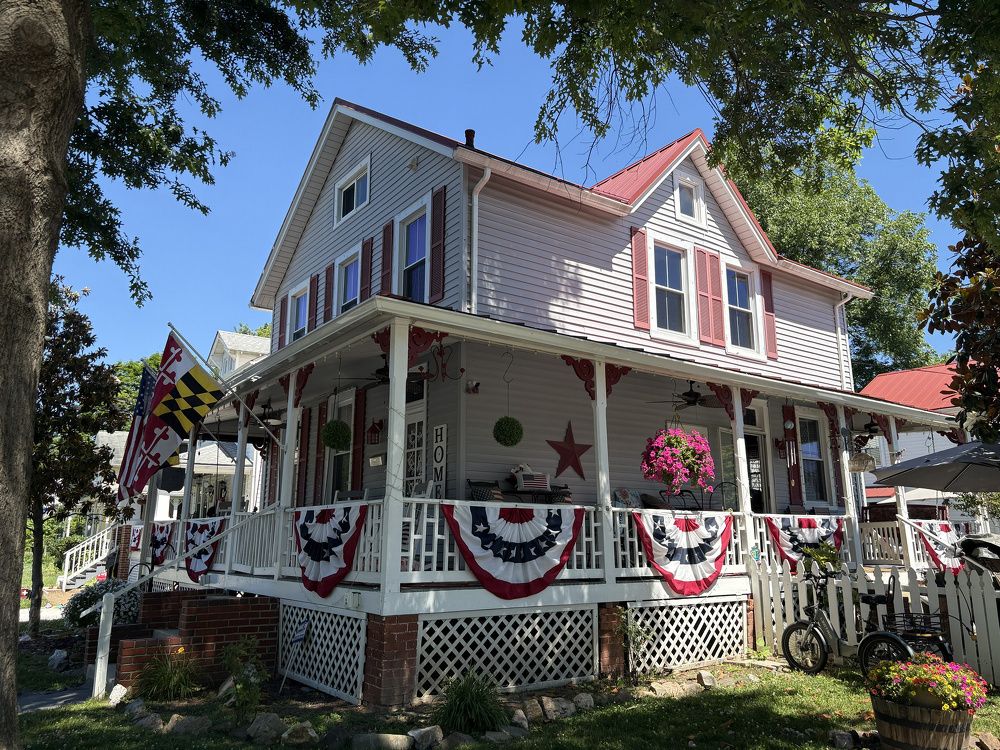 A charming two-story house with a wraparound porch is decorated with patriotic bunting, hanging plants, and flags.