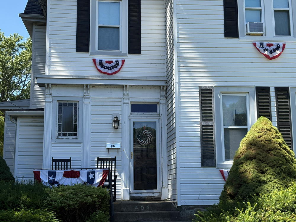 A white two-story house decorated with red, white, and blue patriotic bunting on the porch railing and above the windows.