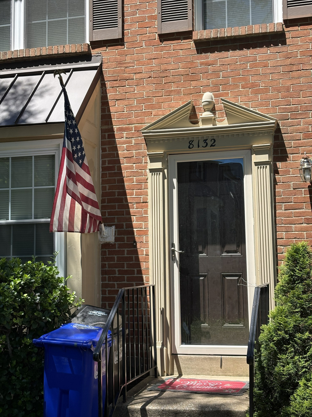 A brick townhouse entrance features a black door with the number 8132 above it, an American flag hanging beside a window, a blue recycling bin, and greenery on either side of the steps.