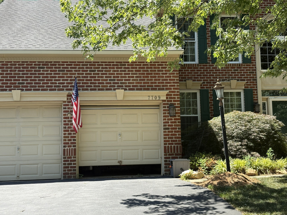A residential brick house features a two-car garage partially open with an American flag mounted on the left side and is surrounded by greenery and a decorative lamp post.