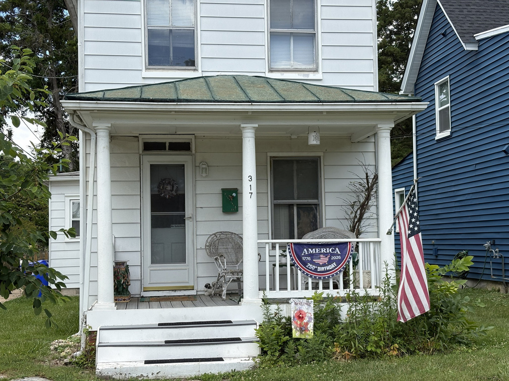 A white two-story house with a small porch featuring a rocking chair, an American flag, and patriotic decorations is shown.