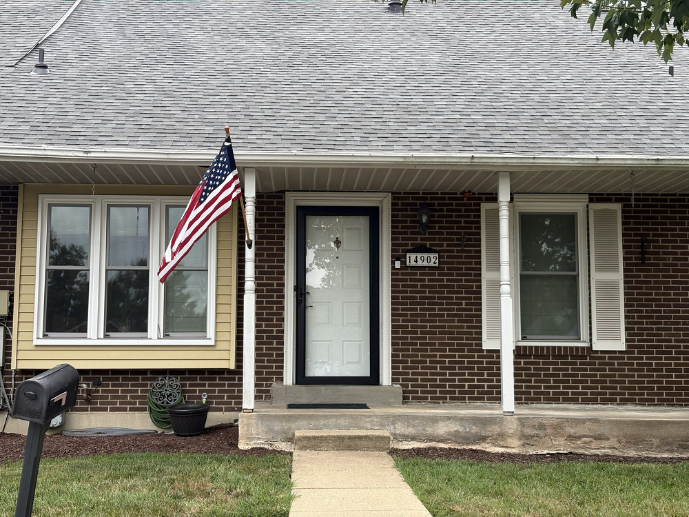 A brick and siding house features a white front door, two windows with shutters, an American flag mounted on a porch column, and a mailbox in the front yard.