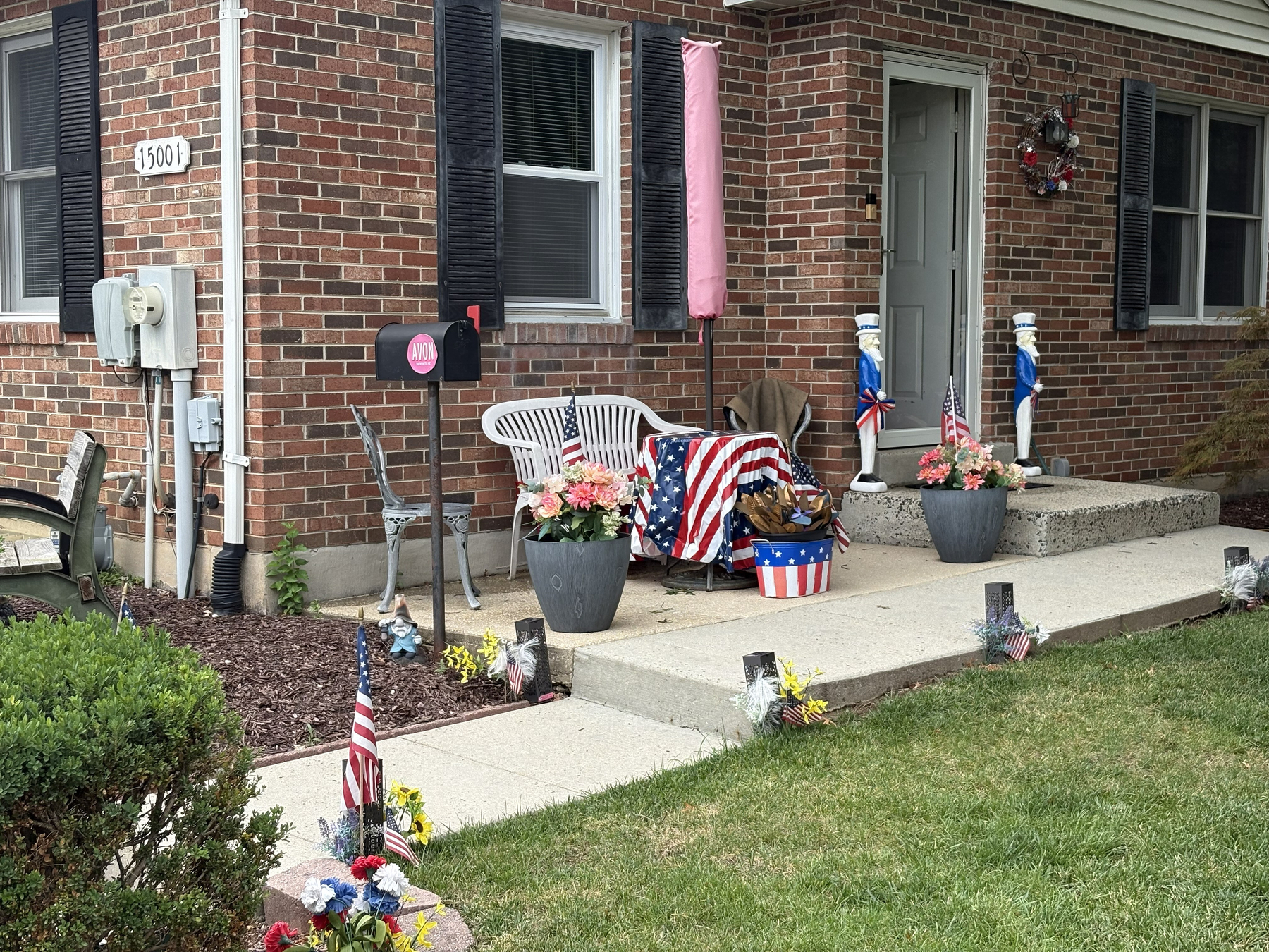 Auto-generated description: A brick house porch decorated with patriotic American flags, flowers, lawn ornaments, and a chair draped with a stars-and-stripes blanket.