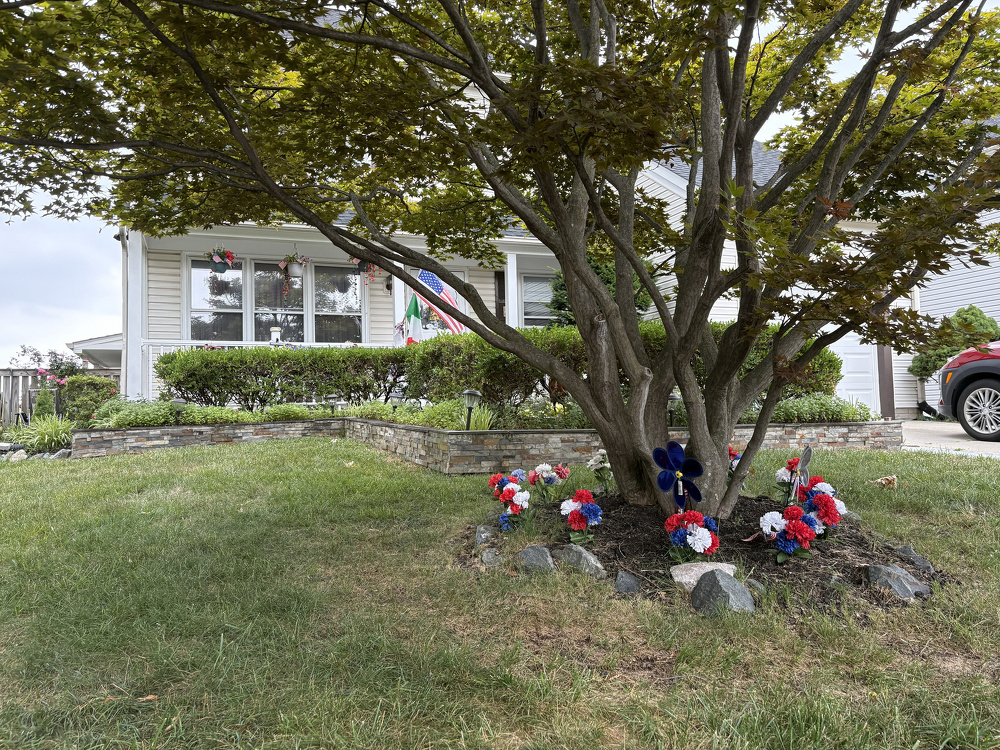 A house with a well-maintained front yard featuring a tree surrounded by red, white, and blue decorative flowers and an American flag hanging by the entrance.