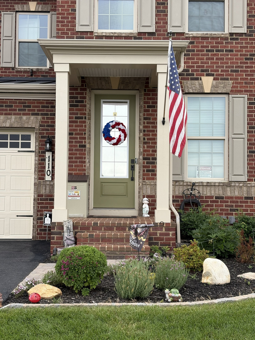A brick house features a green front door decorated with a red, white, and blue wreath, an American flag hanging on the porch, and a small garden with various plants and decorations.