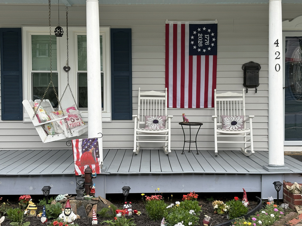 A cozy front porch features swing seats, rocking chairs, an American flag, and a charming flower garden decorated with gnomes.