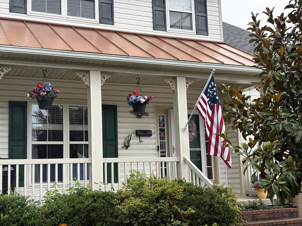 A porch of a house features hanging flower baskets, an American flag, and greenery in front.