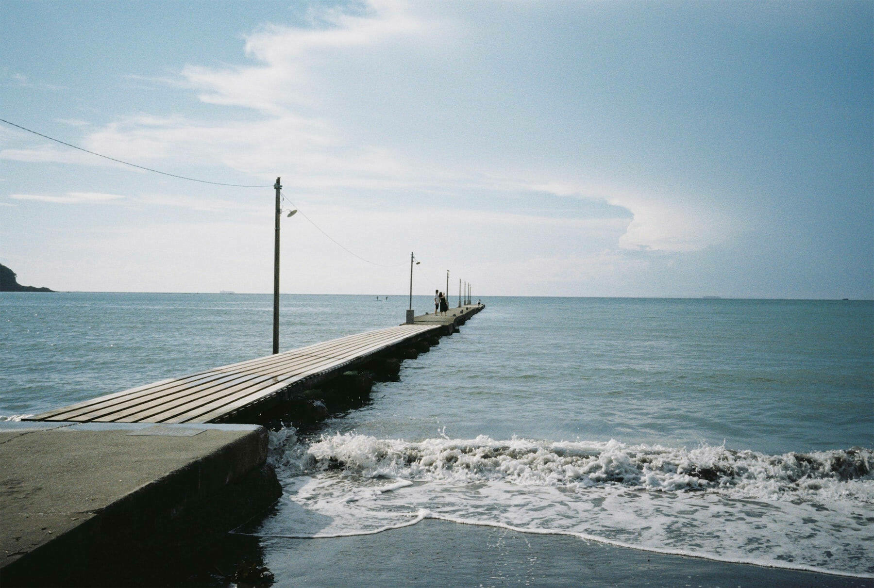 A long wooden pier stretches out into the calm ocean under a partly cloudy sky.