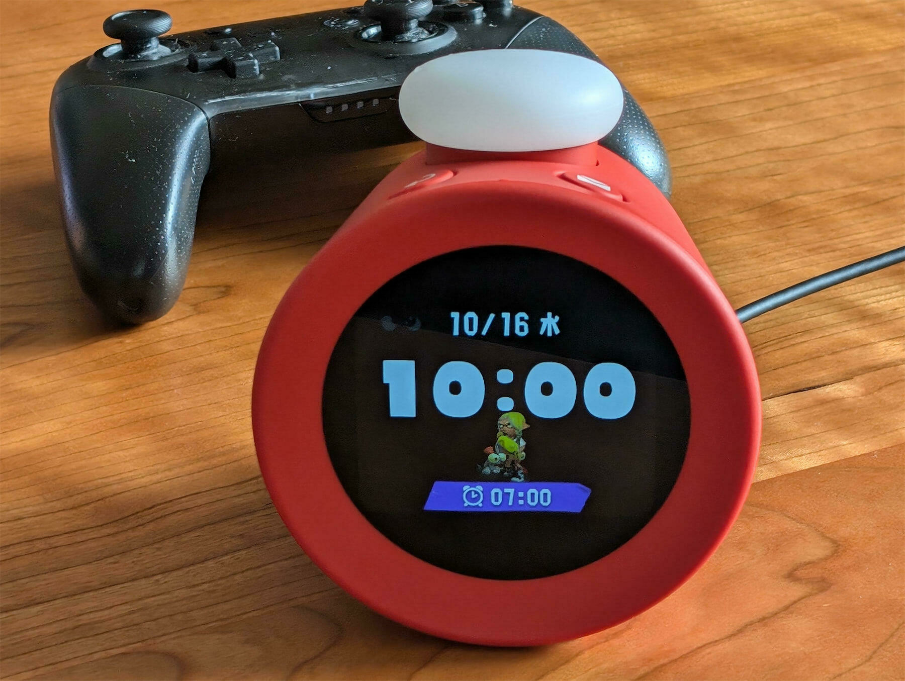 Auto-generated description: A digital alarm clock with a cartoon character displayed is on a wooden surface next to a game controller.