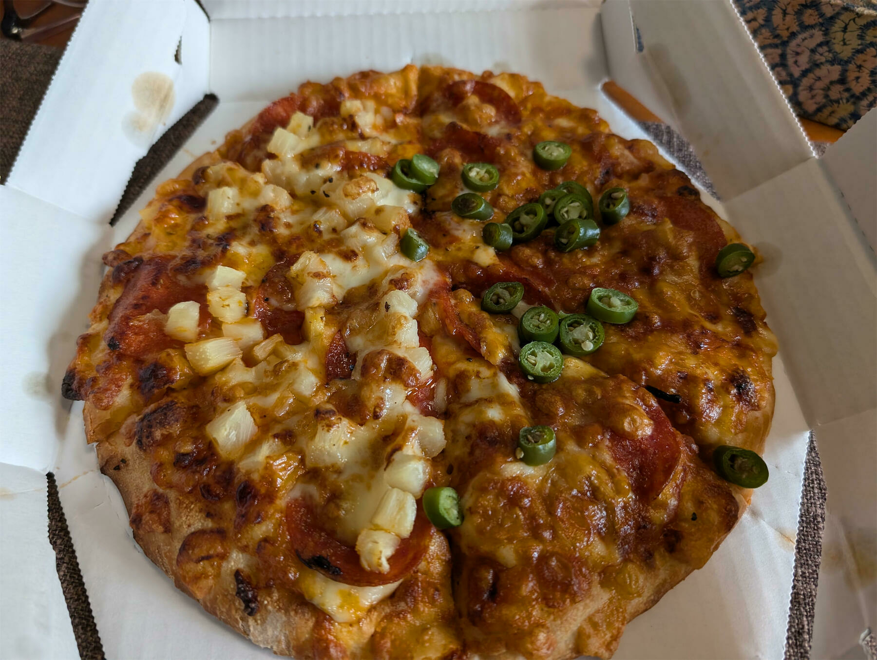 A pizza in a box is topped with melted cheese, diced potatoes, pepperoni, and sliced green peppers.