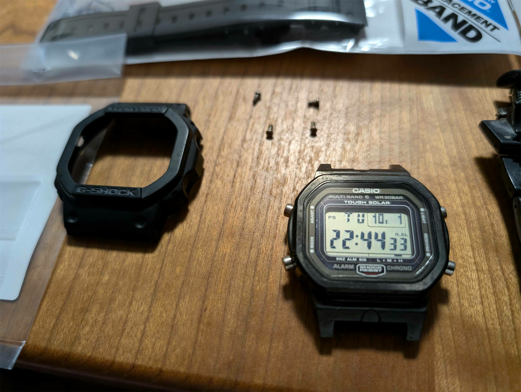 A disassembled Casio GW-5000-1JF watch with its replacement strap and screws laid out on a wooden surface.