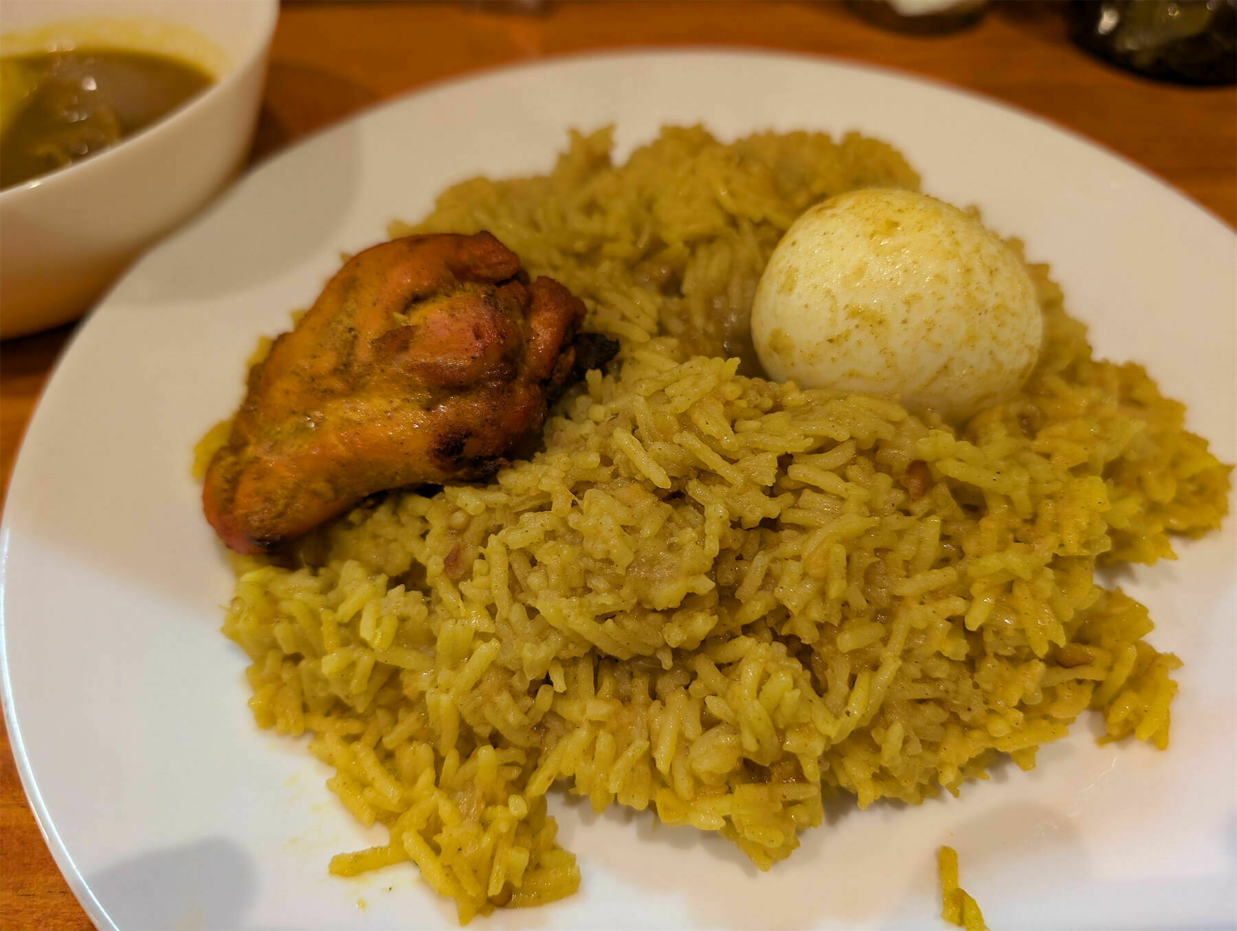 A plate of yellow rice is served with a piece of grilled chicken and a boiled egg, accompanied by a small bowl of sauce.