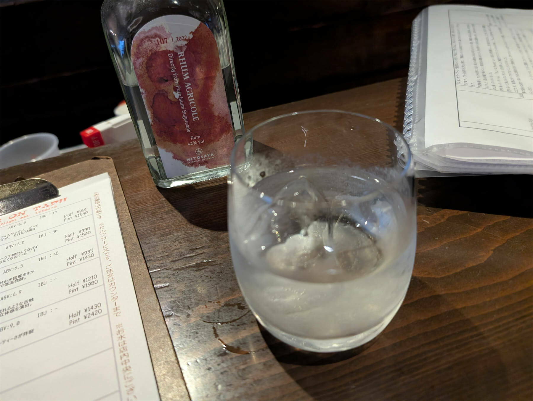 A glass of a clear liquid with ice is placed on a wooden table next to a bottle and some papers.