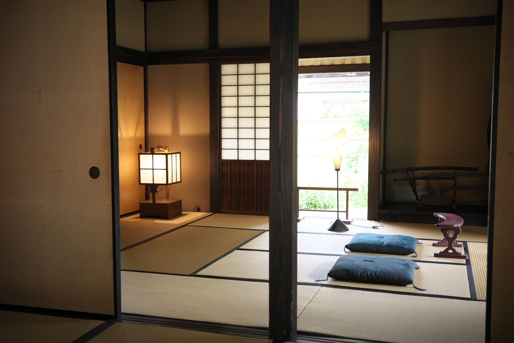 Auto-generated description: A traditional Japanese tatami room features cushions on the floor, a paper lantern, and a view of a garden through a shoji screen.