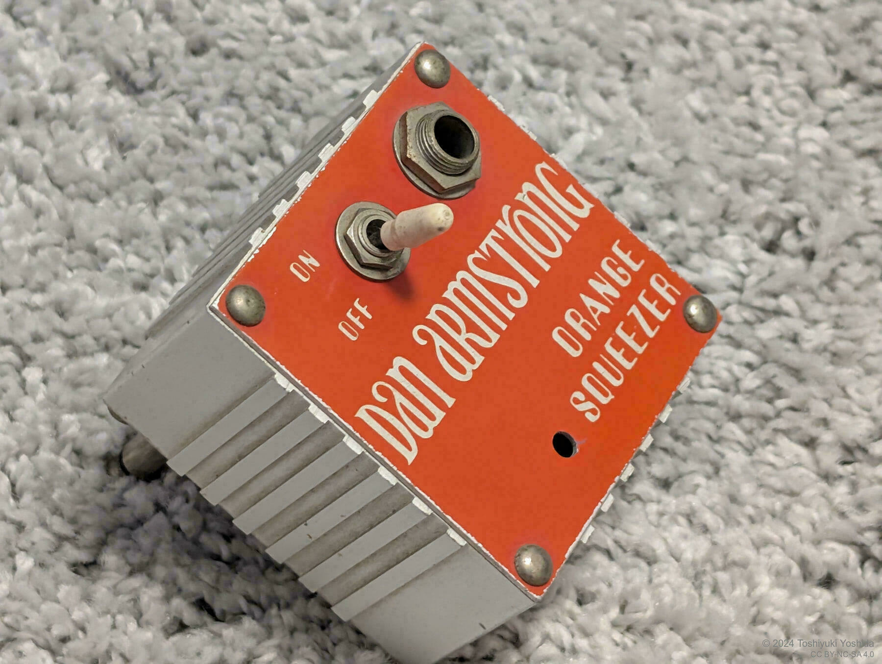 A Dan Armstrong Orange Squeezer guitar effects pedal is placed on a textured surface.