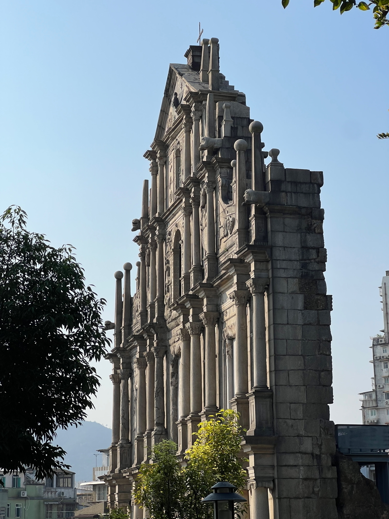 Auto-generated description: The picture shows the ruins of a historic stone facade against a clear blue sky, surrounded by trees and other buildings.