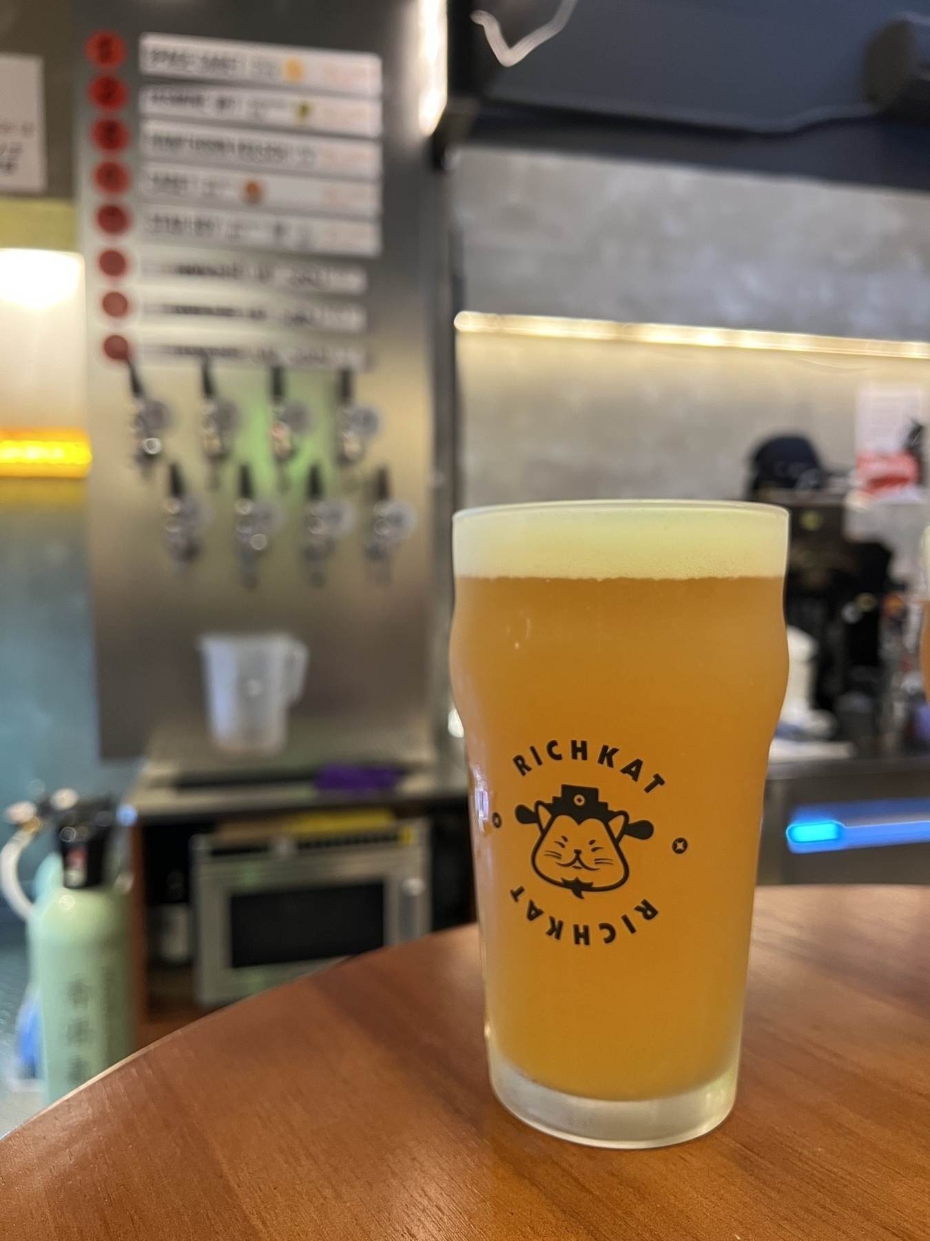 A frothy beer in a glass sits on a wooden table at a bar, with taps and a menu board in the background.