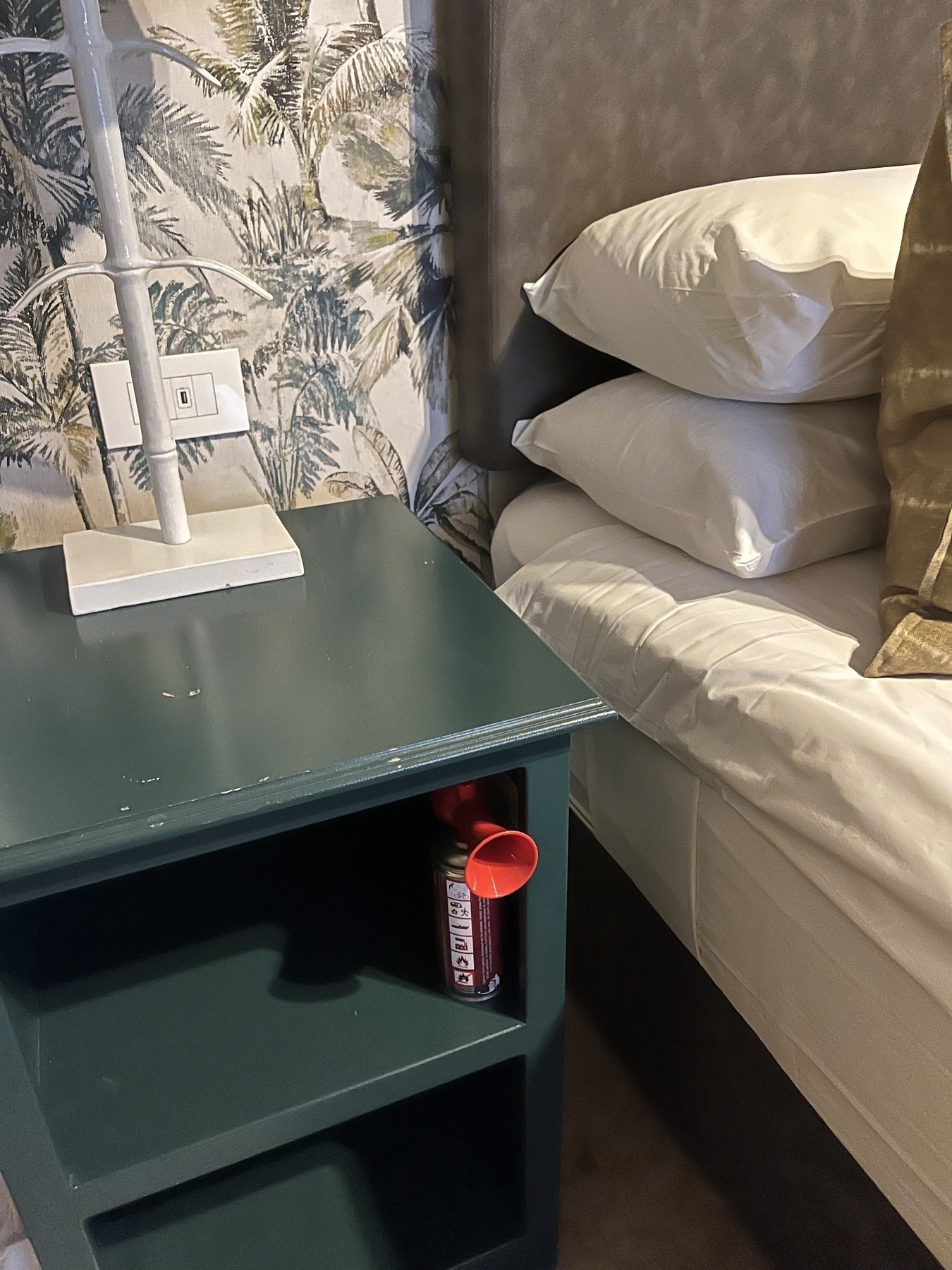 A cozy bedroom corner features a green nightstand with a can of Coca-Cola and a white lamp, next to a neatly made bed with pillows against a patterned wall.