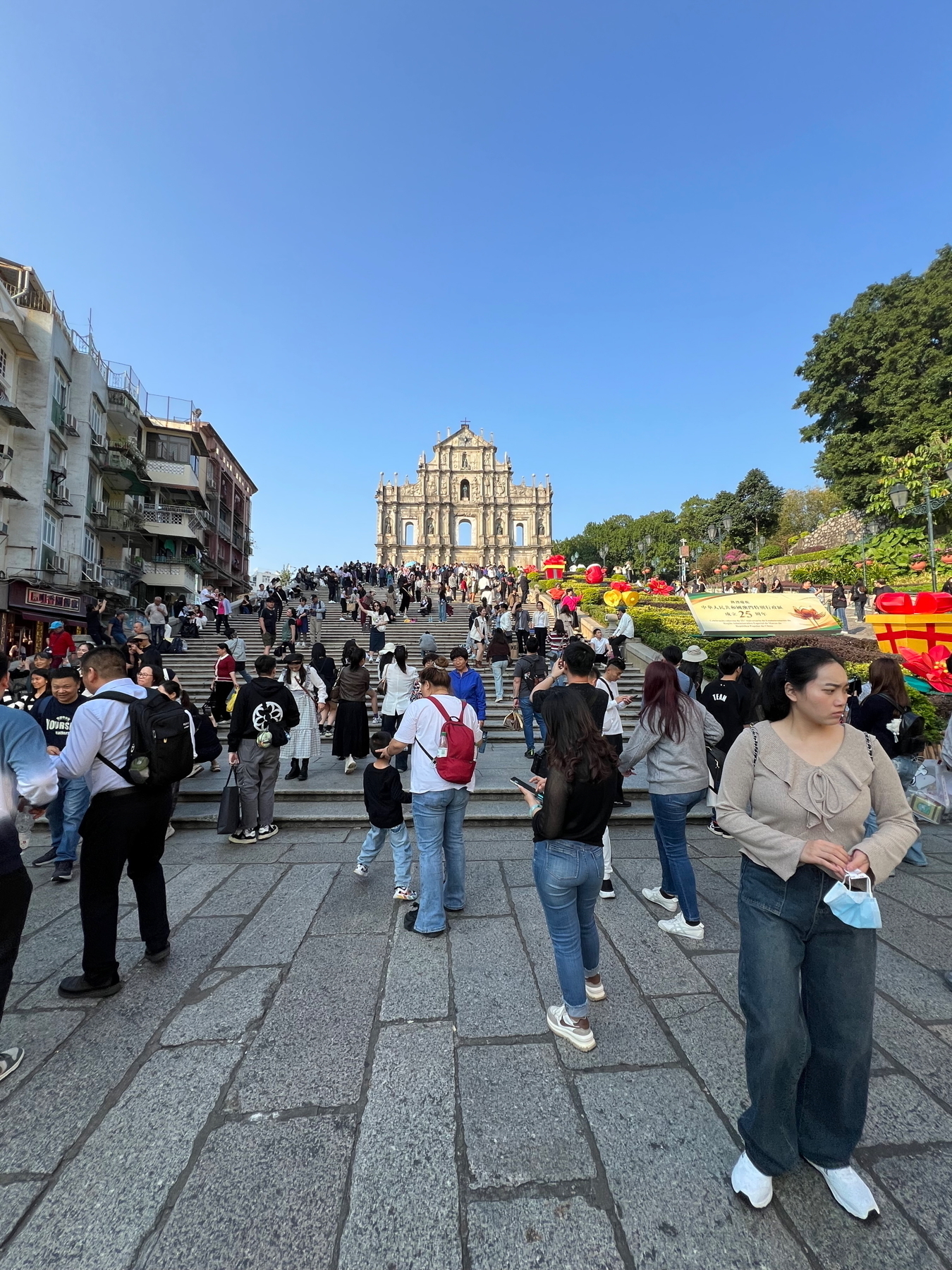 Auto-generated description: A large crowd is gathered in front of the historic ruins of St. Paul's under a clear blue sky.