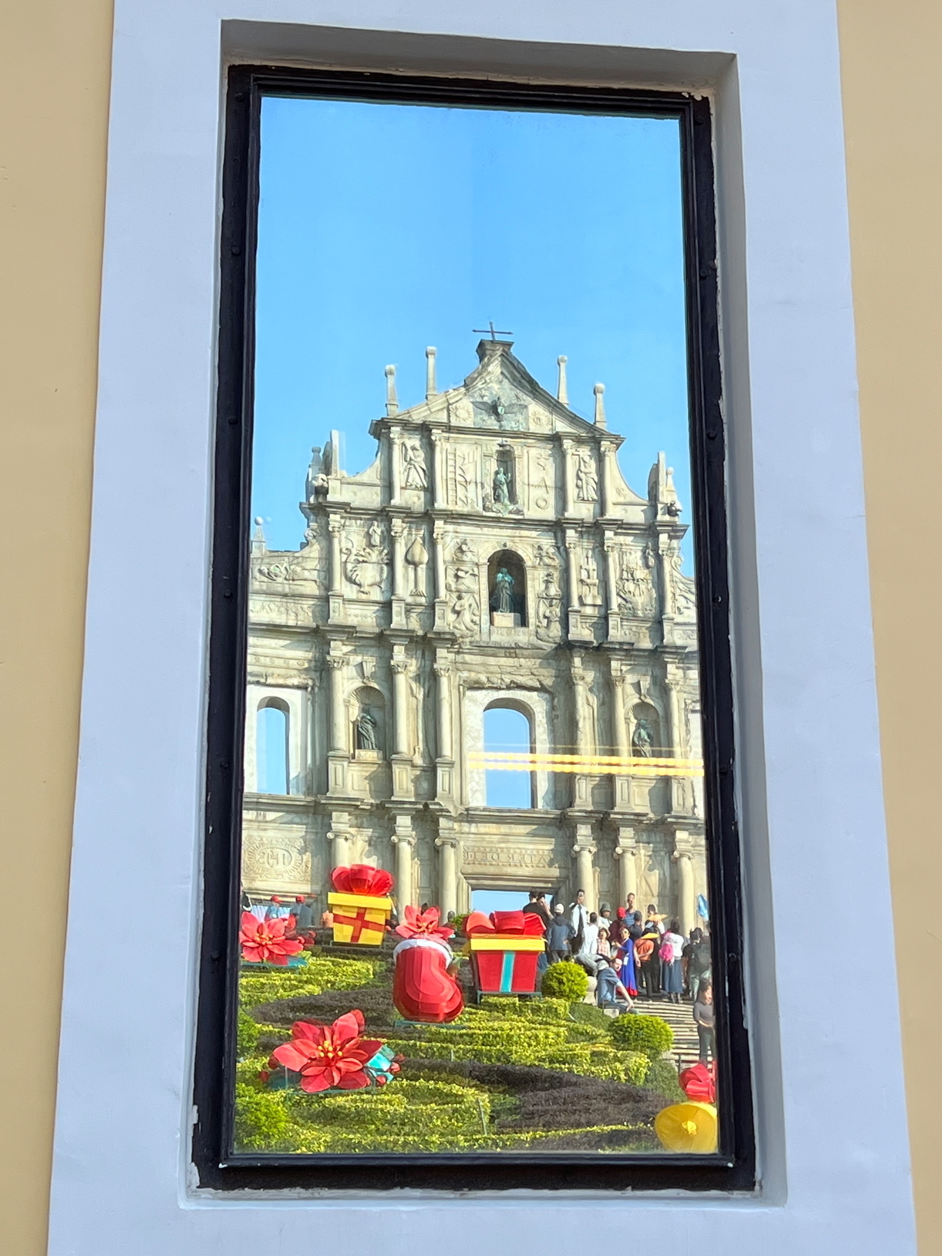 Auto-generated description: A reflection of a historic facade adorned with colorful gift boxes and flowers is framed by a tall window.