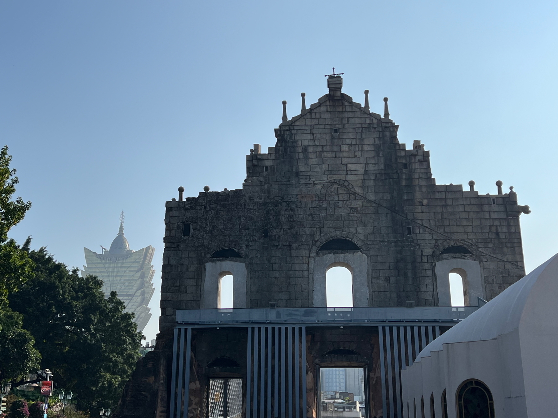 Auto-generated description: Ruins of an old stone facade stand in front of a modern building with a distinctive design under a clear blue sky.