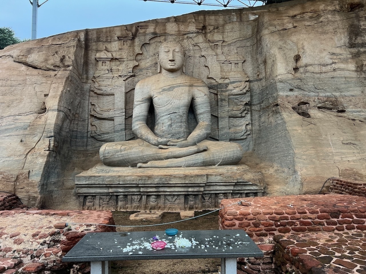 Auto-generated description: A large stone carving of a seated Buddha is embedded in a rocky cliff with offerings placed on a table in the foreground.