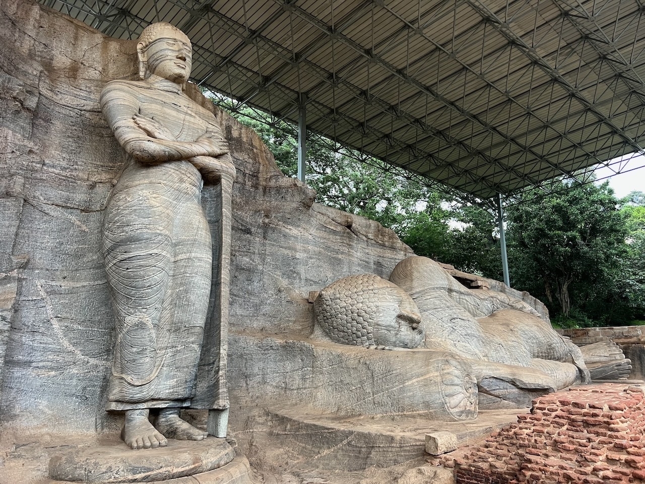 Auto-generated description: A large stone sculpture of Buddha stands next to a reclining Buddha statue carved into a rock face under a protective shelter.