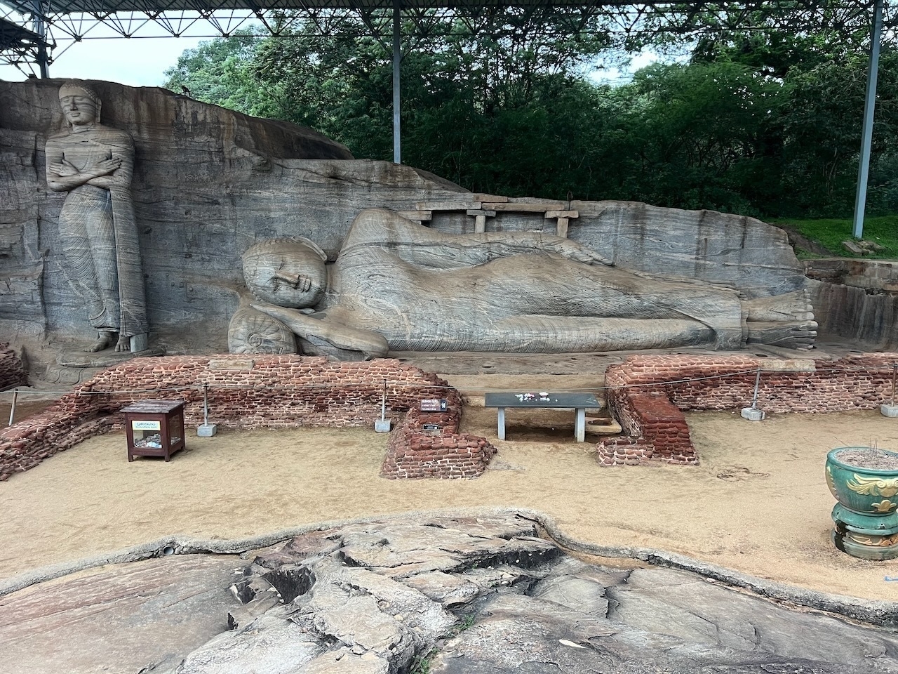 Auto-generated description: A large stone depicts a reclining Buddha, accompanied by a standing figure, set within a historical archaeological site surrounded by lush greenery.