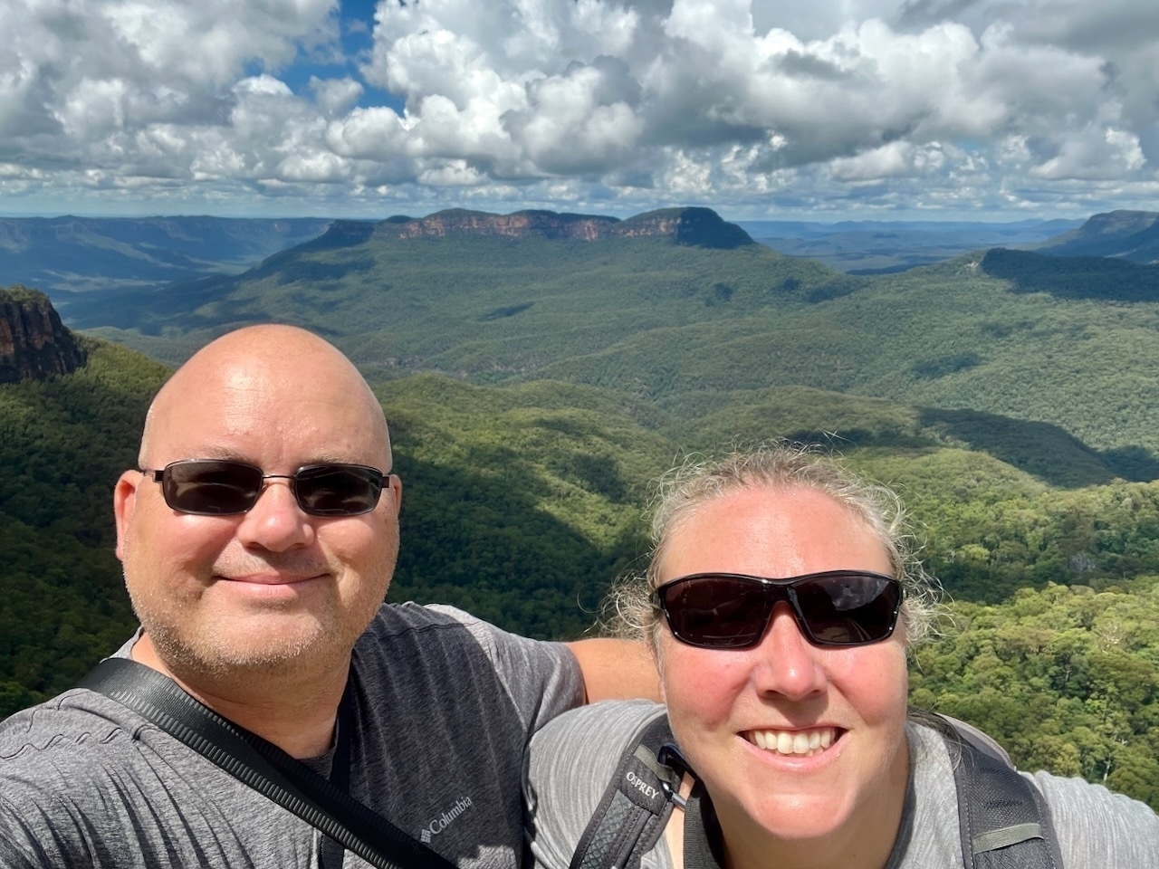 Auto-generated description: Two people wearing sunglasses are smiling for a selfie with a vast, mountainous landscape in the background under a partly cloudy sky.