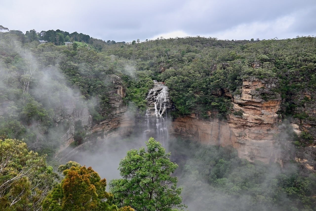 Auto-generated description: A waterfall cascades down a lush, forested cliff surrounded by mist and dense greenery.