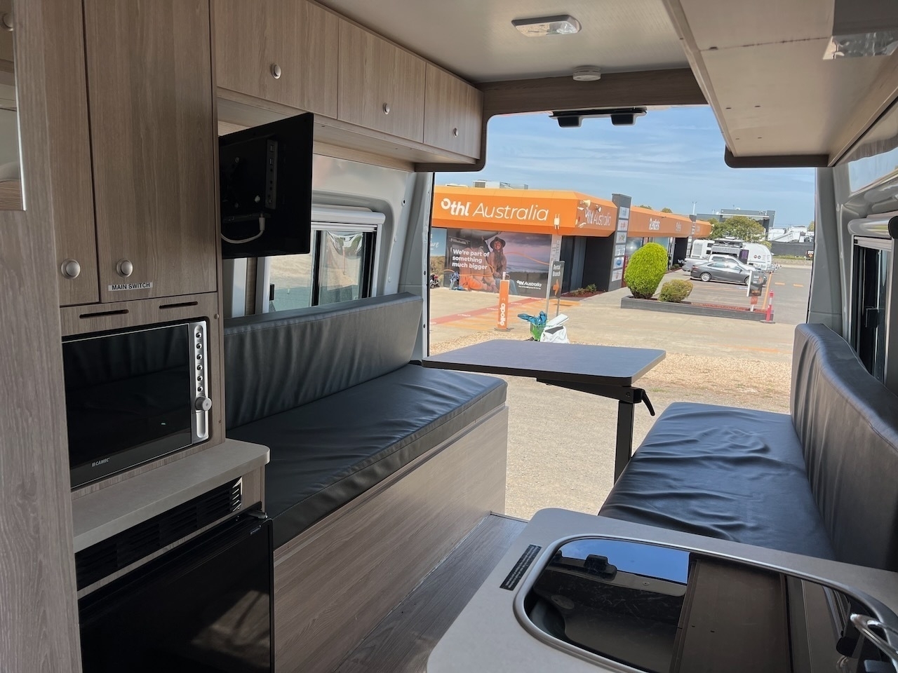 Auto-generated description: A campervan interior features seating, a small table, a sink, and a microwave, with a view of a caravan dealership outside.