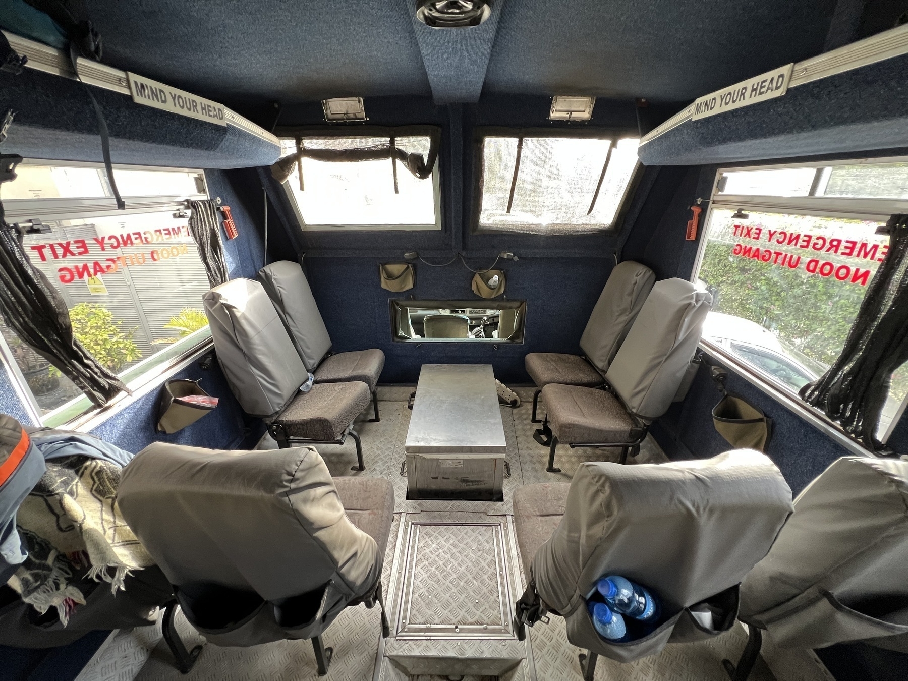 Auto-generated description: A spacious, upholstered vehicle interior with multiple cushioned seats and large windows, featuring safety signs and emergency exits.